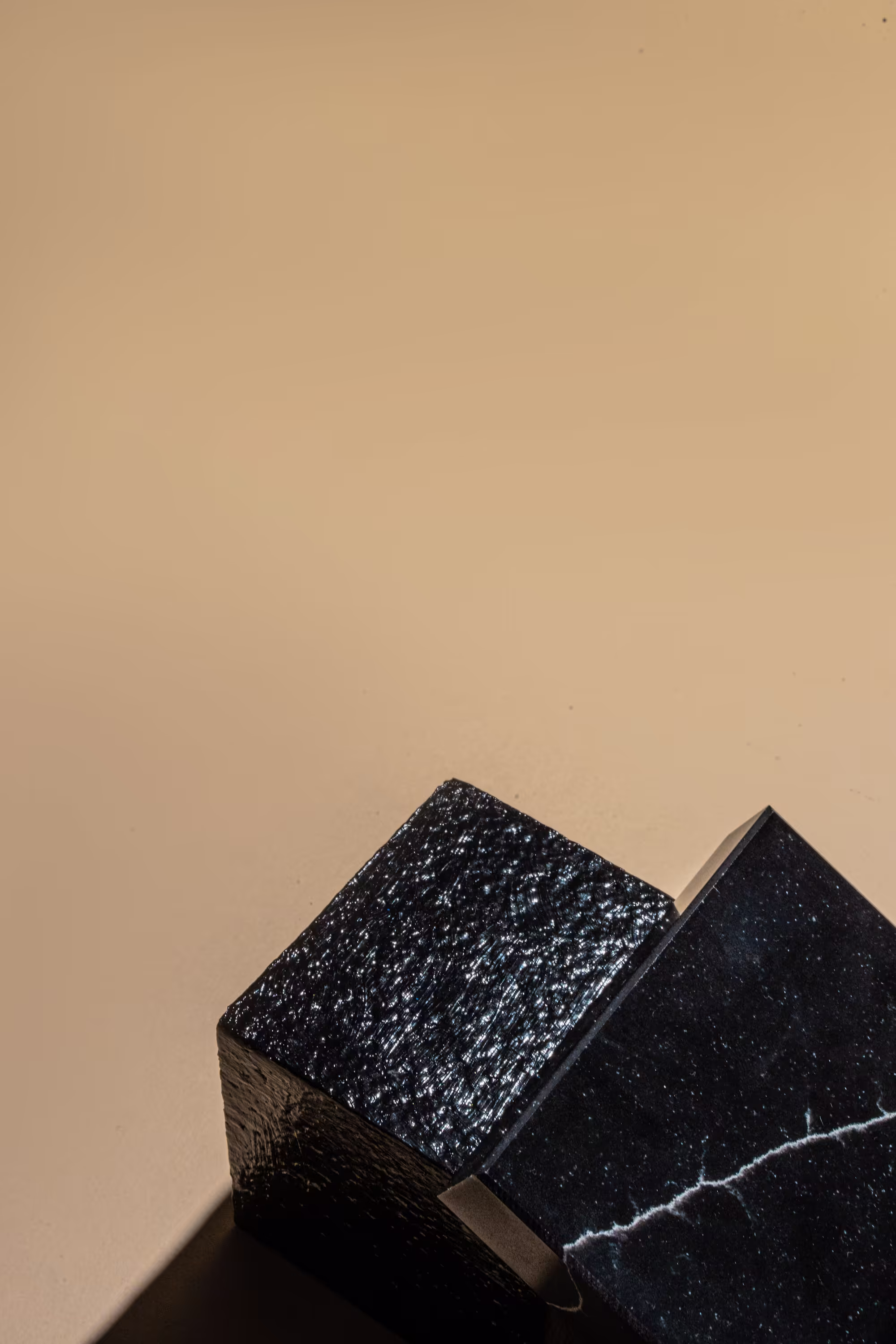 Close-up of two black textured cubes stacked at an angle on a beige surface, one cube has a rough matte finish and the other shows a smooth surface with a white crack pattern.
